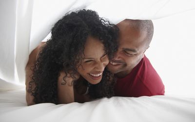 Close up shot of couple laughing, while lying on a bed pulling a sheet over their heads. 